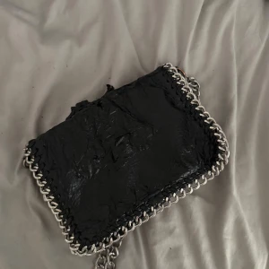 Svart axelväska med kedjedetaljer - Cool svart axelväska med grova silverfärgade kedjor runt kanterna och som axelrem. Väskan har ett lock med magnetknappar och en innerficka med dragkedja. Materialet är skinnliknande och ger en edgy vibe.