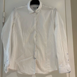 Vit skjorta från J. Harvest & Frost - Klassisk vit skjorta från J. Harvest & Frost i rak modell med lång ärm och knappar framtill. Skjortan har diskreta blå detaljer vid knappslå och manschetter. Perfekt för en clean och stilren look.