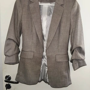 Beige kavaj från H&M, storlek 34 - Stilren beige kavaj från H&M i storlek 34. Kavajen har klassisk krage, två fickor med lock och är tillverkad i en blandning av polyester, viskos och elastan. Fodret är ljusgrått och ger en snygg kontrast. Perfekt för en clean och modern look.