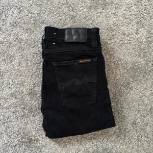 Svarta jeans från Nudie Jeans, 29/34 - Säljer ett par svarta jeans från Nudie Jeans i modellen Tight Long John. Klassisk femficksdesign, svarta sömmar och diskret logga på bakfickan. Jeansen är i stretchig bomull och har dragkedjegylf. Perfekta för dig som gillar en slim och stilren look.