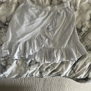 Ljusblå volangkjol - Supersöt ljusblå kortkjol med broderade små blommor och volangdetaljer längs nederkanten och omlott framtill. Kjolen har resår i midjan och är gjord i ett lätt bomullstyg, perfekt för varmare dagar. Ger en romantisk och fräsch vibe till din outfit.