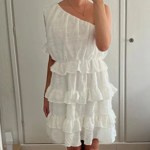 Vit volangklänning  - Superfin vit kortklänning från Dennis Maglic med volanger i flera lager och oneshoulder-design. Klänningen har en luftig känsla och är perfekt för varma dagar. Superfin till student, konfirmation eller skolavslutning. 