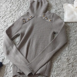 Beige ribbad turtleneck - Säljer en beige ribbad turtleneck med långa ärmar och snygga guldknappar på axlarna. Tröjan är stickad och har en huva som ger en chill vibe. Perfekt för dig som gillar stilrena detaljer och vill ha något bekvämt men ändå trendigt.