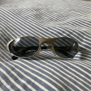 Silvriga sportiga solglasögon Åhus Beach - Snygga solglasögon med sportig vibe från Åhus Beach Handboll Festival. De har ovala, mörka linser och en silvrig båge i syntetmaterial. Perfekta för dig som vill sticka ut med en cool och futuristisk look - väldigt robust material - känns solida. 
