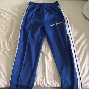 Blå Palm Angels mjukisbyxor med ränder - Snygga blå mjukisbyxor från Palm Angels med vita och svarta ränder längs sidorna. Byxorna har elastisk midja och logga tryckt på ena benet. Perfekta för en chill och sportig look. Bara använda en gång.