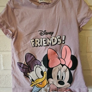 Ljuslila Disney t-shirt H&M 110/116 - Söt ljuslila t-shirt från H&M med Disney-tryck av Kajsa och Mimmi samt texten 'FRIENDS!'. Klassisk rund hals och korta ärmar. Perfekt för alla som älskar Disney och vill ha en färgglad och lekfull stil. Ett litet hål vissa Kajsas rosett som knappt syns.
