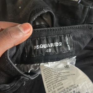 Svarta Dsquared2 jeans med slitningar - Säljer ett par svarta jeans från Dsquared2 i storlek 48. Jeansen har coola slitningar, färgstänk och en tvättad look. Klassisk femficksmodell med Dsquared2-logga vid fickan. Sitter snyggt och har en modern passform.