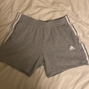 Grå Adidas shorts med vita ränder - Snygga grå shorts från Adidas med klassiska vita ränder längs sidorna och Adidas-logga framtill. Mjukt material och elastisk midja för skön passform. Perfekta för träning eller chill dagar.