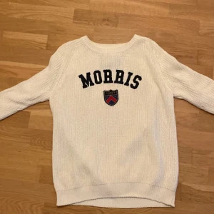 Vit Stickad tröja Morris - Säljer en stilren vit stickad tröja från Morris med text och emblem på framsidan. Tröjan har långa ärmar och en rund halsringning. Perfekt för en klassisk och avslappnad look.