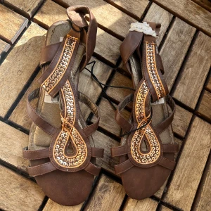 Bruna sandaler med paljettdetaljer - Snygga bruna sandaler från Vox Shoes med remmar och glittrande paljettdetaljer i guld längs ovansidan. De har öppen tå och platt sula, perfekt för varma dagar. Unik design med dekorativa mönster framtill som ger extra bling till outfiten.