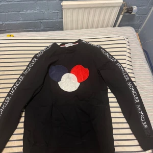Svart Moncler sweatshirt med logga - Svart sweatshirt från Moncler med lång ärm och tryckt Moncler-logga i blått, vitt och rött på bröstet. Moncler-tryck längs båda ärmarna ger en sportig vibe. Skön passform och klassisk rund halsringning. Använd få gånger  Storleken är XL men passar M den är lite i storlek   Köpt på vinted
