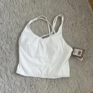 Vit linnetopp från Aim'n med smala band - Supersnygg vit linnetopp från Aim'n med tunna axelband som korsas i ryggen. Toppen är croppad och har en stilren design med diskret Aim'n-logga baktill. Perfekt för träning eller chill dagar, tillverkad i mjukt och stretchigt syntetmaterial. Prislapp kvar, säljer då den är för stor för mig