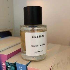 ESSNCE Sweet Cara Eau de Parfum - En stilren parfymflaska från ESSNCE med doften Sweet Cara. Flaskan är cylindrisk i genomskinligt glas med svart rund kork och vit etikett med minimalistisk text. Innehållet är ljusgult och parfymen är en Eau de Parfum. Använd litegrann. 