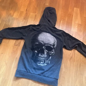 Grå hoodie med dödskalle från Philipp Plein - Säljer en mörkgrå hoodie från Philipp Plein med stor dödskalle-tryck på ryggen och svarta detaljer längs ärmarna. Dragkedja framtill, huva och diskret logga på bröstet.