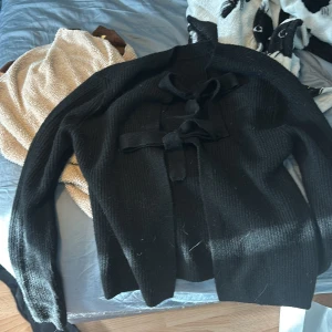 Svart stickad kofta med rosett från Pieces - Svart stickad kofta från Pieces i storlek XS. Koftan har långa ärmar och stängs framtill med två dekorativa rosetter. Den är ribbstickad och har en klassisk, enkel design som är lätt att matcha med olika outfits.ANDVÄND EN GÅNG!!