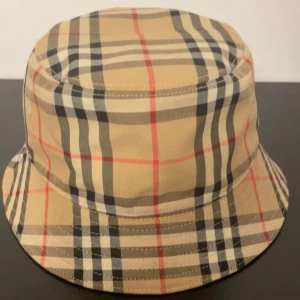 Ekta burberry hat  - Bucket hat från Burberry i klassiskt beige rutigt mönster med svarta, vita och röda linjer. Hatten har en mjuk, rund form och är tillverkad i bomull. Perfekt accessoar för att lyfta din stil med en ikonisk touch. Med Box