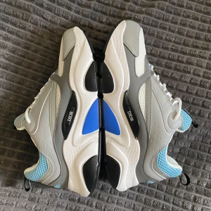 Chunky D sneakers i grått och blått - Säljer ett par chunky D sneakers med cool design i grått, vitt och ljusblått. Skorna har meshdetaljer, skinnpaneler och vit sula med svarta och blå inslag. Tjock plattform och snörning framtill. Perfekta för dig som vill sticka ut med lyxig streetstyle.