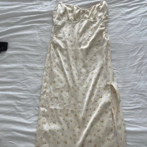 Ljusblommig långklänning med slits - Superfin långklänning i ljus satinliknande material med små gula blommor. Klänningen har smala axelband, söt snörning vid bysten och en snygg slits på sidan. 