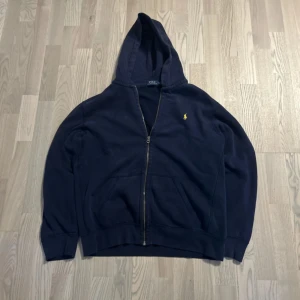 Marinblå hoodie från Polo Ralph Lauren XL - Snygg marinblå hoodie från Polo Ralph Lauren i storlek XL. Klassisk design med dragkedja, huva och känguruficka. Broderad gul logga på bröstet. Perfekt för chill dagar eller när du vill ha en stilren streetlook.