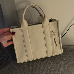 Beige handväska i skinn med dragkedja - Snygg beige handväska i stilren boxig modell med två handtag och praktisk dragkedja upptill. Väskan har flera fack och är gjord i ett slätt skinnmaterial. Perfekt storlek för det viktigaste och passar till många outfits.