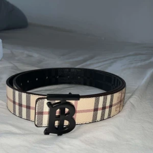 Burberry vändbart bälte med B-spänne - Snyggt vändbart bälte från Burberry med klassiskt rutigt mönster på ena sidan i beige, svart och rött, och svart rutigt mönster på andra sidan. Stort svart B-spänne framtill. Perfekt accessoar för att lyfta din outfit.