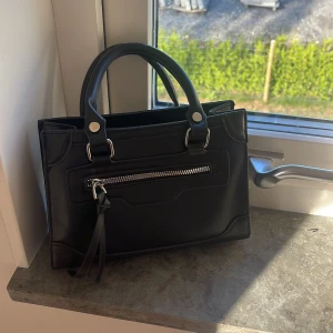 Svart liten handväska  - Snygg svart handväska i skinn med stilrena silverdetaljer och praktiskt ytterfack med dragkedja framtill. Väskan har två handtag och ett stort utrymme inuti. Perfekt storlek för det viktigaste och snyggt. 
