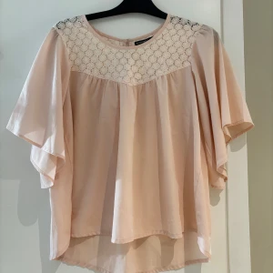 Ljusrosa blus med spets från Rut&Circle - Supersöt ljusrosa blus från Rut&Circle med vida, fladdriga ärmar och vacker vit spets upptill. Blusen har rund halsringning och knapp i nacken. Perfekt för dig som gillar romantiska detaljer och en luftig känsla.