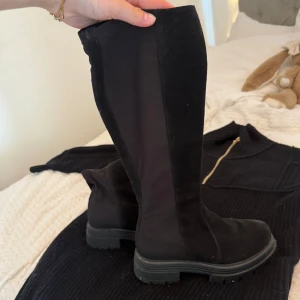Svarta höga boots med chunky sula - Snygga svarta höga boots med chunky platt sula och rund tå. Ovandelen är i en mix av mocka och stretchigt syntetmaterial som sitter tight längs benet. Perfekta för att lyfta din outfit med en edgy vibe.🖤😍🌟