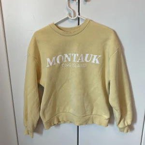 Gul sweatshirt Montauk Gina Tricot XS - Gul sweatshirt från Gina Tricot med vit text 'MONTAUK LONG ISLAND' framtill. Klassisk rund halsringning, ribbade muddar och avslappnad passform. Perfekt för chill dagar och enkel att styla med jeans eller leggings. Väl använd men bra skick!