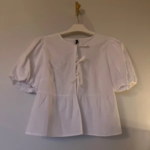 Vit blus med puffärm och knytningar - Supersöt vit blus med puffiga korta ärmar och tre knyt framtill. Blusen är lite kortare och har en lätt volang nedtill. Oanvänd, endast testad🤍