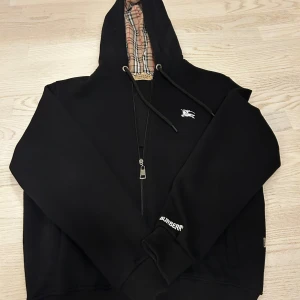 Svart Burberry hoodie zip *kvitto medföljer* - Svart hoodie från Burberry med klassiskt rutigt foder i huvan och broderad logga på bröstet. Tröjan har dragkedja framtill, snörning i huvan och ribbade muddar. Broderad Burberry-text på ärmen ger extra premiumkänsla. 
