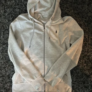 Ljusgrå hoodie från Polo Ralph Lauren - Snygg ljusgrå hoodie från Polo Ralph Lauren med dragkedja och huva. Klassisk logga broderad på bröstet, snörning i huvan och två fickor framtill. Perfekt för en chill och stilren look.