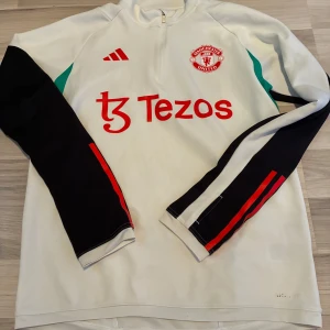 Manchester United träningströja Adidas M - Snygg vit Manchester United träningströja från Adidas med svarta ärmar, röda detaljer och gröna inslag på sidorna. Halv dragkedja framtill, långärmad och slim fit. Perfekt för fotbollsträning eller när du vill representera ditt lag.