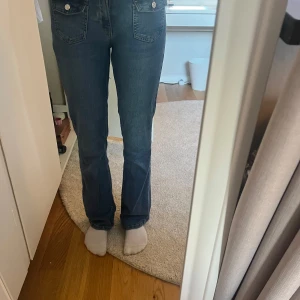 Blå bootcut jeans med medel midja - Säljer ett par blå jeans med bootcut-modell och medelhög midja. Jeansen har framfickor med lock och knappar, klassisk femficksdesign och är tillverkade i ett mjukt denimtyg.