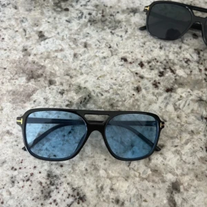 Svarta Tom Ford solglasögon med blått glas - Snygga svarta solglasögon från Tom Ford med blåtonade glas och klassisk pilotform. Ramen är i svart plast med guldiga T-detaljer på sidorna och överdelen. Perfekt accessoar för att lyfta din stil och sticka ut i mängden. (R3plica)