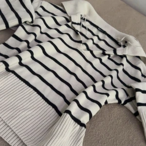 Randig stickad tröja från Gina Tricot - Vit och svart randig stickad tröja från Gina Tricot med krage och v-ringning. Tröjan har långa ärmar och breda ribbade muddar vid ärmslut och nederkant. Perfekt för en chill och stilren look.