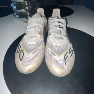 Adidas F50 vita fotbollsskor - Säljer ett par Adidas F50 PRO Road to Glory fotbollsskor i vitt med svarta och lila detaljer samt guldiga inslag på sulan. Skorna har snörning, klassiska tre svarta ränder på sidan och mönstrad sula med dobbar för gräsplan. Lätta och snygga skor för dig som vill sticka ut på planen. Skorna köptes för 1 månad sedan och är knappt använda 9/10 skick. Hör av vid funderingar