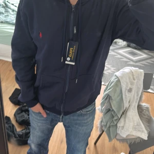 Marinblå hoodie från Polo Ralph Lauren - Snygg marinblå hoodie från Polo Ralph Lauren med dragkedja och huva. Klassisk röd logga broderad på bröstet och praktiska fickor framtill, perfekt för chill dagar eller när du vill ha en clean streetstyle.(vet ej om den är äkta)