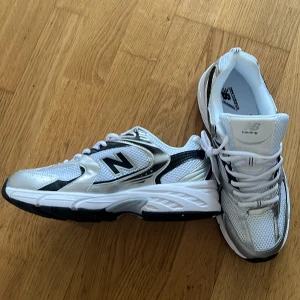 Vita och silver New Balance sneakers - Snygga New Balance sneakers i vitt och silver med svarta detaljer. Skorna har meshpaneler för extra andningsförmåga, klassisk snörning och en chunky sula. Perfekta för dig som gillar sportig och trendig stil. Ikonisk N-logga på sidan och bekväm passform. Oanvänd skor!!
