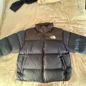 Blå dunjacka från The North Face - Säljer en blå och svart dunjacka från The North Face med hög krage och dragkedja framtill. Jackan har två sidofickor med dragkedja, justerbar nederkant och logga på bröstet och ryggen. Perfekt för kalla dagar och riktigt snygg streetstil.