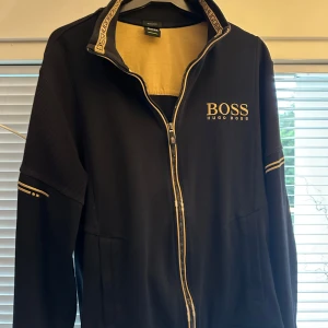 Svart zipjacka från Hugo Boss - Svart zipjacka från Hugo Boss med guldiga detaljer och logga på bröstet. Jackan har hel dragkedja, ribbade muddar och diskreta ränder på ärmarna. Snygg och stilren design med bekväm passform, perfekt för dig som gillar klassisk streetstyle.