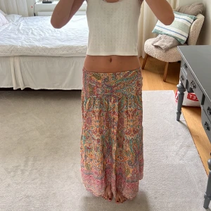 Färgglad långkjol med blommigt mönster - Säljer en superfin långkjol med färgstarkt blommigt mönster i rosa, orange, blått och grönt. Kjolen har en bohemisk vibe med smock i midjan och flowigt fall, perfekt för varma dagar. Materialet känns lätt och luftigt, vilket gör den skön att bära hela sommaren.