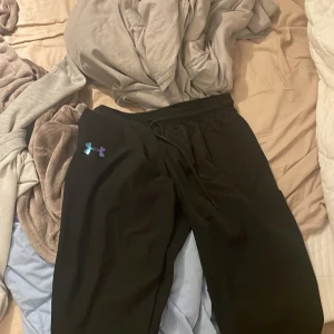 Svarta mjukisbyxor från Under Armour - Svarta mjukisbyxor från Under Armour med dragsko i midjan och stort reflekterande logotryck längs ena benet. Byxorna har även en liten logga på höften och är gjorda i ett mjukt och bekvämt material, perfekta för chill eller träning.Passar även s