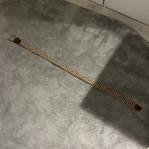 Brunt och beige flätat bälte - Snyggt flätat bälte i brunt och beige med silverfärgat spänne. Bältet har detaljer i brunt skinn vid spännet och ändarna. Perfekt accessoar för att lyfta din outfit med en retrovibe och coolt mönster.