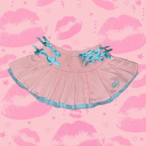 Rosa kjol Dolls Kill x Hatsune Miku - Supersöt limited edition slutsåld rosa kortkjol från Dolls Kill x Hatsune Miku med blåa banddetaljer och broderad logga. Kjolen har plisserad design, blå kant nertill och knytband i midjan. Perfekt för dig som älskar kawaii och statement-plagg!