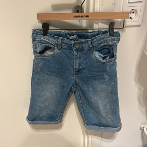 Blå jeansshorts från Name It - Snygga blå jeansshorts från Name It med klassisk femficksdesign, uppvikta benslut och slitna detaljer. Perfekta för en avslappnad stil under varma dagar. Mjuk denim med justerbar midja för extra komfort.