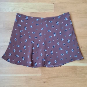 Brun blommig kort kjol från H&M  - Supersöt brun kort kjol från H&M Divided med små blå och vita blommor över hela tyget. Kjolen har en lätt utsvängd form och är gjord i ett mjukt, tunt material som känns skönt mot huden. Perfekt att styla med en enkel topp och sneakers.