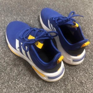 Blå sneakers Adidas strl 38 - Snygga blå sneakers från Adidas med vita ränder på sidorna och gula detaljer på plösen och hälen. Ovandelen är i mesh för bra andningsförmåga och sulan är vit med cloudfoam. Storlek 38 (5 1/2)