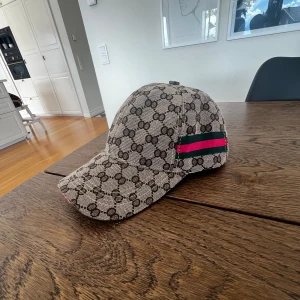 Gucci keps  - Snygg keps från Gucci med klassiskt GG-monogram i brunt på beige botten. Kepsen har gröna och rosa ränder på sidorna. Perfekt accessoar för dig som vill sticka ut med en lyxig streetstyle-look.