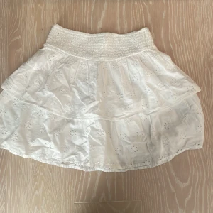 Vit broderad kortkjol från Gina Tricot - Supersöt vit kortkjol från Gina Tricot med bred smock i midjan och broderade blommor över hela kjolen. Kjolen är luftig och har volangig känsla, perfekt för varma dagar. Tillverkad i bomull för en skön och lätt känsla.